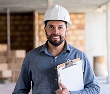 A man wearing a hard hat and a blue shirt is holding a clipboard. He appears to be an archi team member at a construction site.