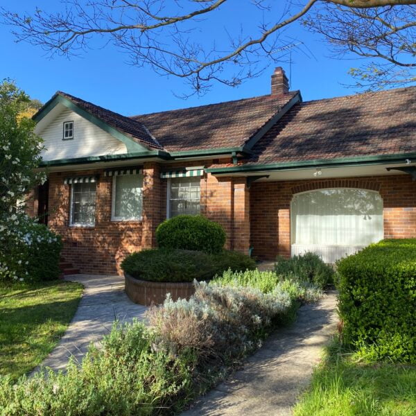A brick house with a tiled roof, chimney, and attached garage. It is surrounded by well-maintained shrubs and greenery, and there is a concrete pathway leading to the front door. Despite its charm, it stands subject to potential home demolitions in the neighborhood.