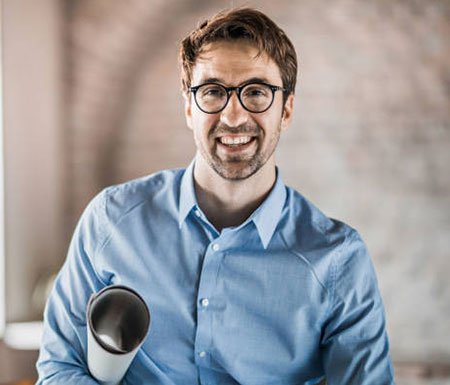 A Member Archi 3, wearing glasses and a light blue shirt, is smiling while holding a metal object.