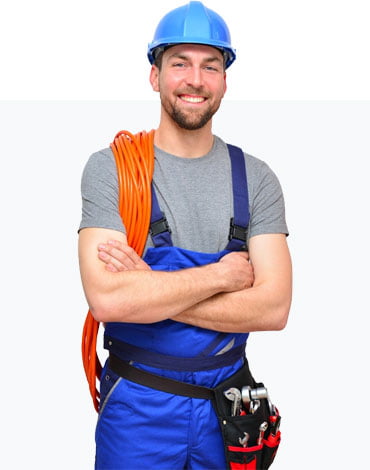 A construction member in blue overalls and a blue hard hat smiles while holding an orange cable over his shoulder and standing with his arms crossed. He has a tool belt with various tools around his waist.