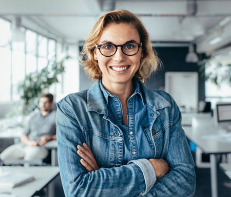 A Member Archi 3 with shoulder-length hair, wearing glasses and a denim jacket, stands smiling with arms crossed in an office setting.