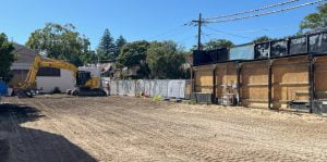 A construction site in Rose Bay with freshly leveled ground, an excavator parked on the left, and a fence separating the site from neighboring houses and structures. Trees and clear blue sky in the background hint at recent demolition in this picturesque Sydney suburb.