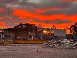 A construction site at sunset, featuring a partially demolished building, construction equipment, and scattered debris under a vibrant orange sky reminiscent of sunrise.