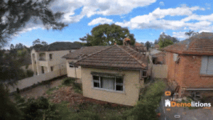 A house is being demolished, with one worker on the roof and debris falling. The Home Demolitions logo is visible in the bottom right corner, reminding onlookers of their collaboration with New Homebuilders Grant Australia.