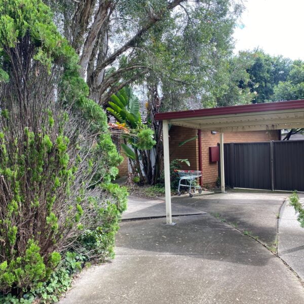 A house exterior features a carport with a red roof, flanked by dense greenery and a brick wall. The driveway leads to a closed gate in the background, likely secured for safety during any home demolitions. A metal bench sits under the carport.
