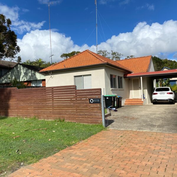 A small single-story house with a red-tiled roof, a wooden fence, a brick driveway, and a white car parked under a carport. The front yard has grass, and trees are visible in the background; though whispers of home demolitions loom over the peaceful neighborhood.