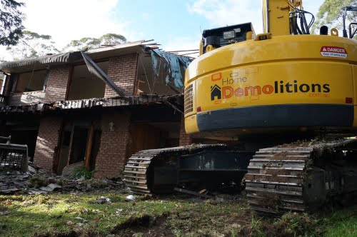 A yellow construction vehicle with the text "Home Demolitions" is parked next to a partially demolished two-story brick house. Debris from the residential demolition is scattered around the machine.
