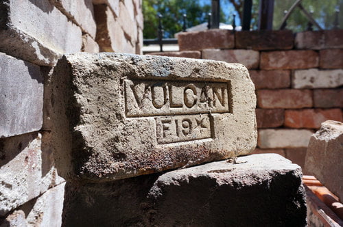 A weathered brick with the inscription "VULCAN F.19X," possibly salvaged from a recent demolition, is surrounded by other bricks in a construction setting, hinting at its former life in a sturdy home.