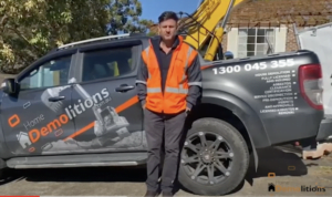 A director in an orange safety vest stands in front of a pickup truck with "Home Demolitions" branding. The truck displays a phone number (1300 045 355) and an excavator arm is visible behind them, sending a clear message of their demolition expertise.