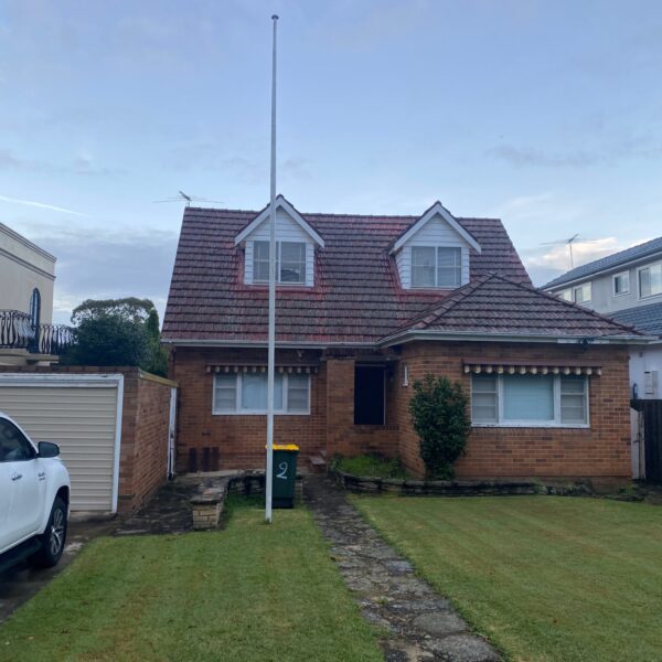 A two-story brick house with a gabled roof and dormer windows. There is a garage to the left, a white truck parked in the driveway, and a flagpole in the front yard. Despite pending home demolitions in the area, there’s still pride on display with a green bin near the entrance.