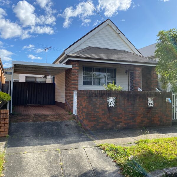 A brick house with a front yard and carport, enclosed by a wrought iron fence. The house has a gable roof and a small window, while the driveway is made of brick pavers. Considering its condition, it might soon face home demolitions for redevelopment.