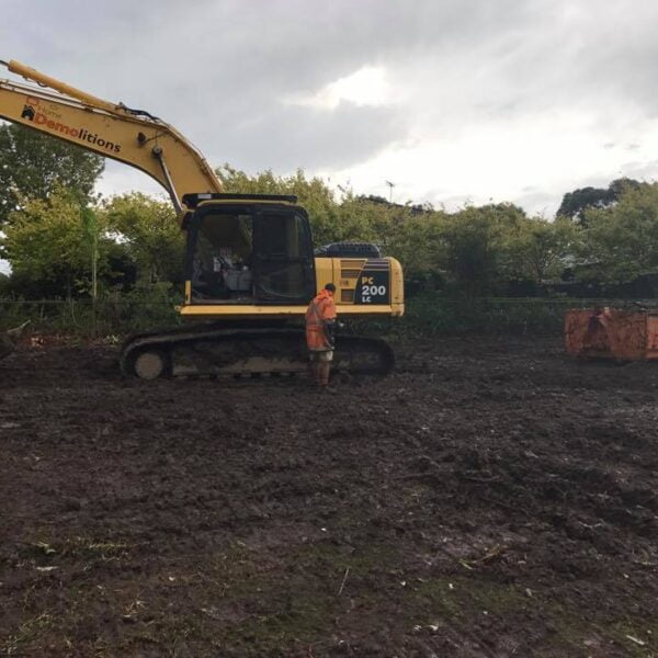 A person in orange workwear stands beside a large yellow excavator on muddy ground, ready for home demolitions, with an orange container nearby and trees in the background.