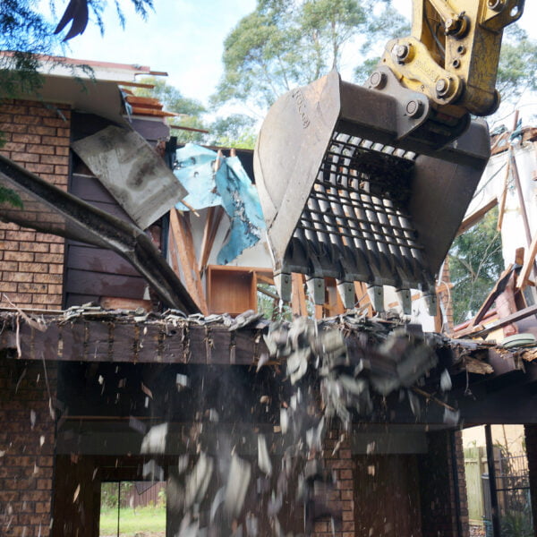 A mechanical excavator demolishes the upper portion of a brick house during home demolitions, causing debris to fall.