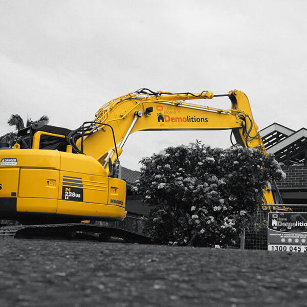 A yellow excavator with "Home Demolitions" on the arm is positioned in front of a partially demolished brick building, with a contractor sign displaying "Demolitions" and a phone number in the foreground.