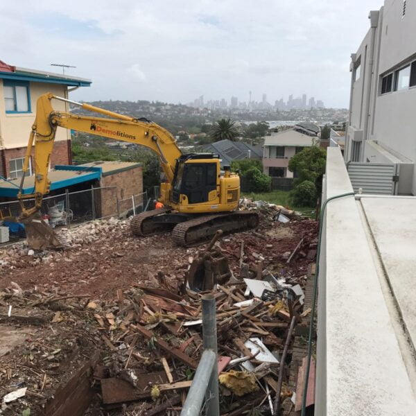 A yellow excavator is clearing debris on a construction site between two buildings, likely from recent home demolitions. Rubble and broken materials are scattered on the ground, while the city skyline looms in the foggy background.