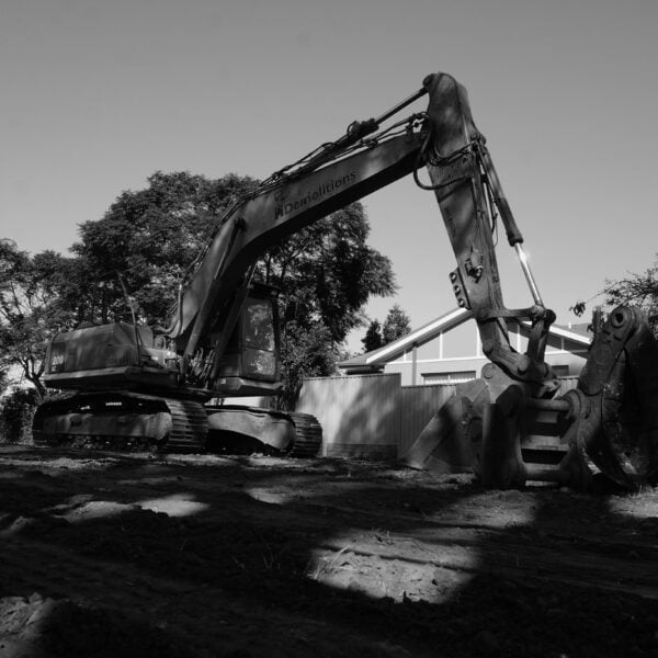 A large excavator sits idle on a construction site, ready for home demolitions, with trees and a building in the background. The image is in black and white.