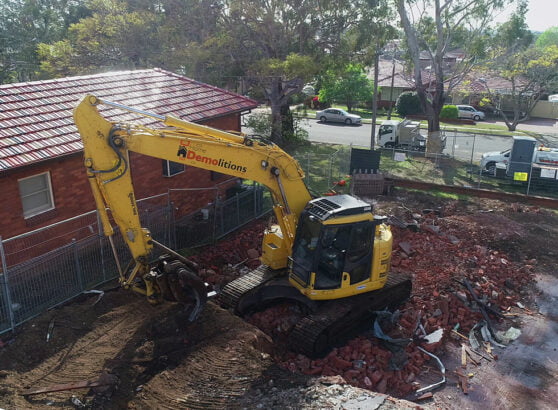 A yellow excavator is engaged in the home demolition process of a red-brick building, with rubble scattered around the site and houses in the background.