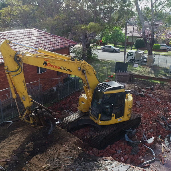 Miranda, NSW 2228 2 A yellow excavator is engaged in the home demolition process of a red-brick building, with rubble scattered around the site and houses in the background.