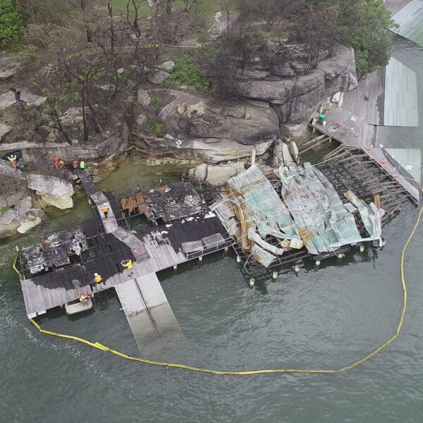 Longueville, NSW 2066 1 Aerial view of a partially burnt dock beside a rocky shoreline. Burnt wooden structures and debris from recent home demolitions are visible on the dock, with a barrier placed around the area in the water. Several people are on-site.