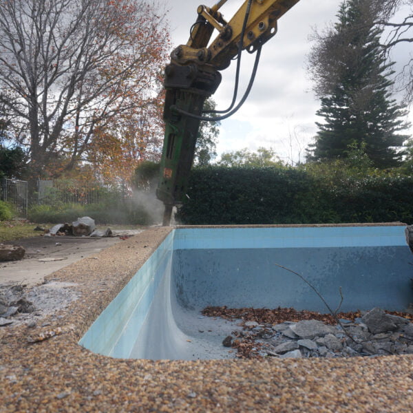 An excavator from the home demolitions crew is hard at work, demolishing a backyard swimming pool surrounded by debris and autumn trees.