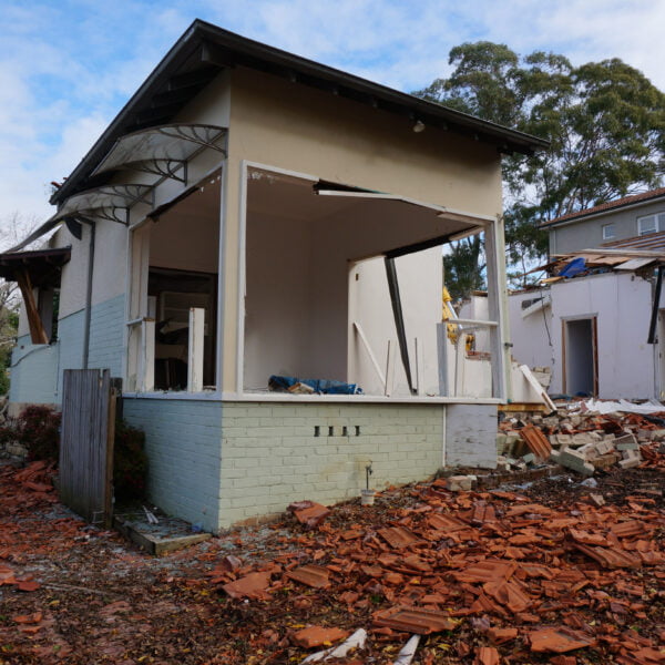 Turramurra, NSW 2074 18 Partially demolished house with scattered bricks and debris in the foreground, illustrating the aftermath of home demolitions. A section of the neighboring house is also visible. Trees and a cloudy sky form the backdrop.