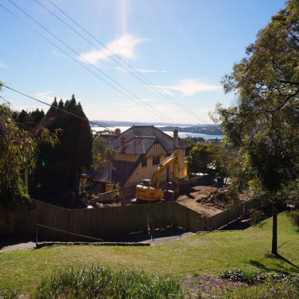 Bellevue Hill, NSW 2023 17 A residential construction site with a yellow excavator, surrounded by trees and houses, hinting at ongoing home demolitions. Power lines are visible against the clear sky, with a distant view of a body of water in the background.