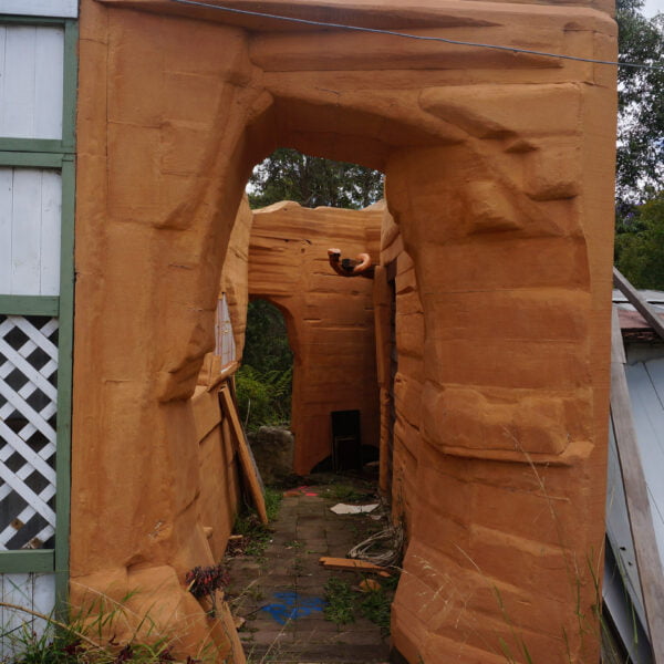 Penrith, NSW, 2750 13 A partially constructed or abandoned building with a faux rocky facade and a lattice wall section on the left stands amid the remnants of home demolitions. Debris is scattered around the ground while vegetation is visible in the background, adding to the scene's desolate charm.
