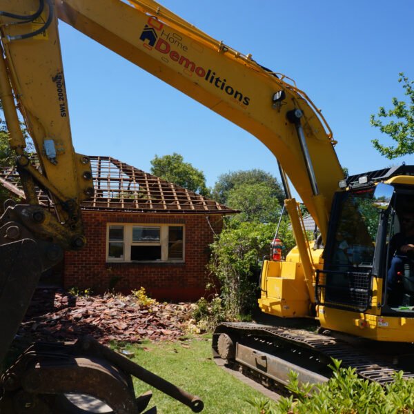 Saint Ives, NSW 2075 12 A yellow excavator is performing home demolitions on a brick house, with part of the roof already removed. The scene is set in a residential area with trees in the background.