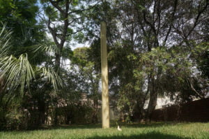 A tall, rectangular monolith stands in the middle of a grassy area surrounded by trees, reminiscent of a site untouched by home demolitions.