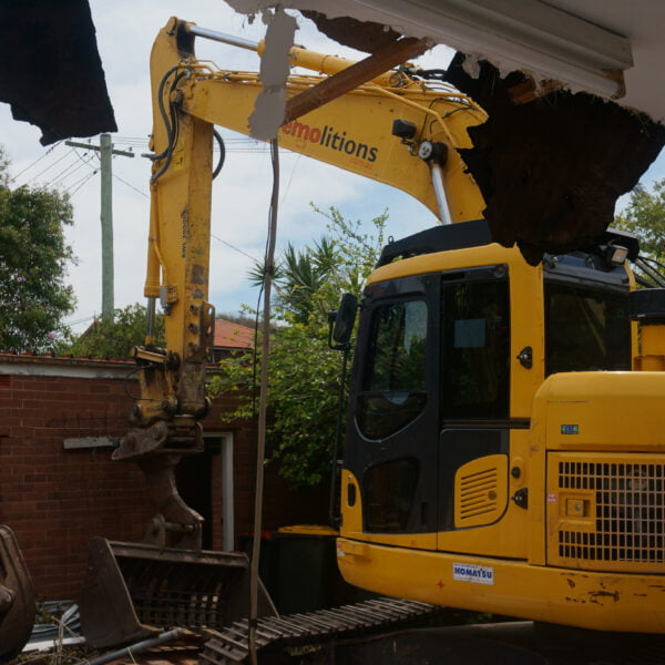 Rodd Point, NSW 2046 10 A yellow excavator labeled "Home Demolitions" demolishes part of a brick building's roof. Trees and power lines are visible in the background.
