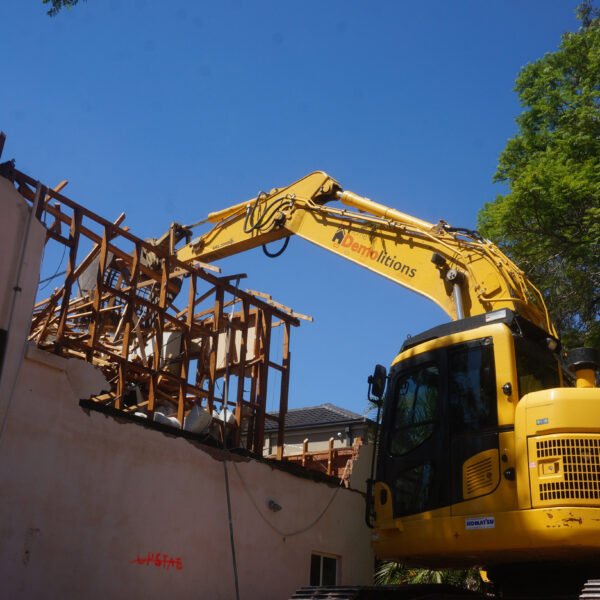Strathfield, NSW 2135 9 A yellow excavator is conducting home demolitions, tearing down the upper structure of a pink building and removing wooden beams and debris. Trees stand tall under a clear blue sky in the background.