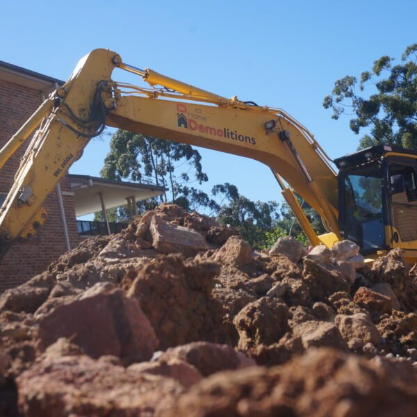 A yellow excavator labeled "Q & J Demolitions" expertly moves debris near a brick building on a sunny day, with trees in the background, showcasing their proficiency in home demolitions.