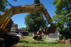 A large yellow excavator with the branding "Home Demolitions" is seen in a grassy yard, operated by a person in an orange vest and helmet. Debris and metal fencing are visible in the background.