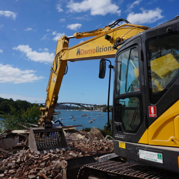 Henley, NSW 2111 7 A yellow excavator from a home demolitions company is working amidst rubble, with a body of water and a bridge visible in the background on a clear day.