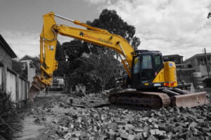 A yellow excavator with "Home Demolitions" on the arm is parked on a pile of rubble at a construction site surrounded by trees and buildings.