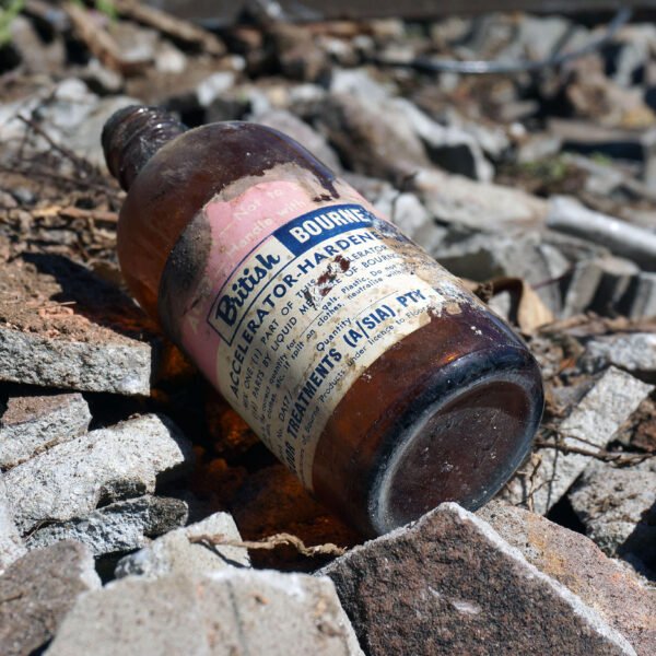 A vintage glass bottle labeled "Betaine and Butyl Alcohol," partially buried in a pile of broken concrete debris outdoors, appears to be a remnant from old home demolitions.