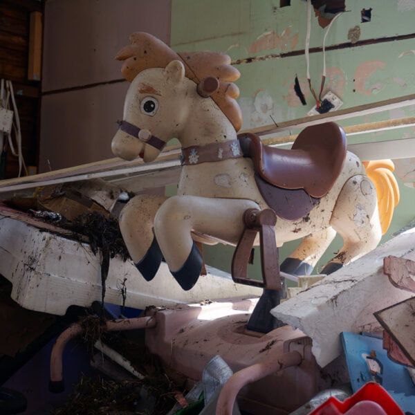 An abandoned, weathered children's mechanical horse ride sits among debris and broken furniture in a dilapidated room, with peeling paint and exposed wires—a haunting remnant of home demolitions.