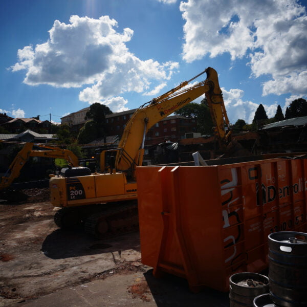 Two yellow excavators work at a home demolition site under a partly cloudy sky. An orange demolition container is in the foreground, with various debris spread around.