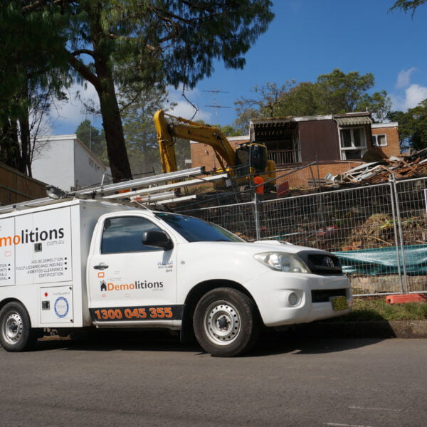 A white truck labeled "Home Demolitions" is parked on the street in front of a fenced demolition site, where an excavator from Home Demolitions is tearing down a residential building.