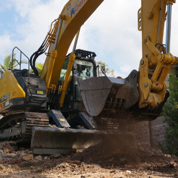 A large, yellow excavator moves a bucket of soil and debris at a construction site. Trees and a house are visible in the background, amidst preparations for home demolitions.