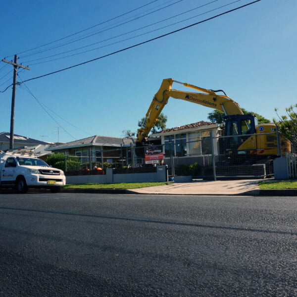 An excavator and a white van are parked outside a single-story house undergoing home demolition on a sunny day.
