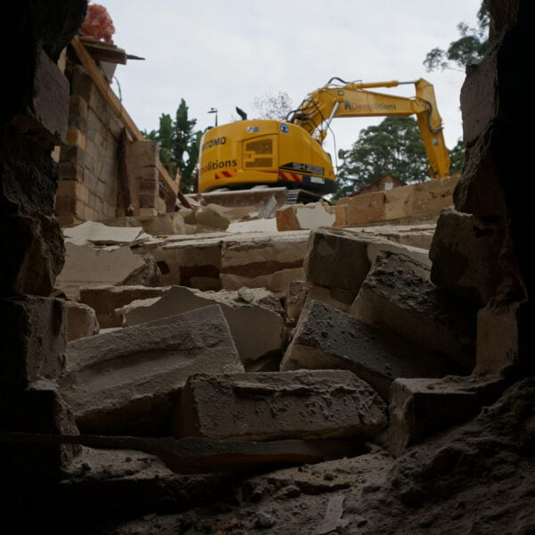 View through a hole in a brick wall showing rubble and a yellow excavator working on home demolitions in the background.