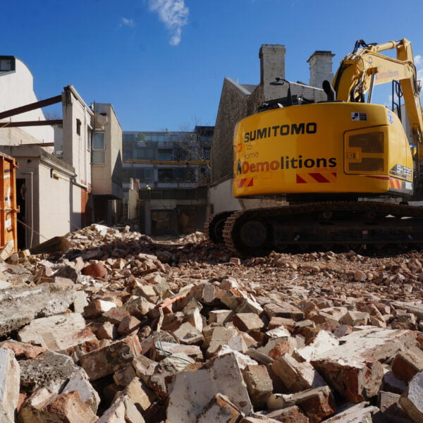 A demolition site with scattered rubble, a large excavator labeled "Sumitomo," and partially demolished buildings in the background under a clear blue sky, showcasing the intense process of home demolitions.