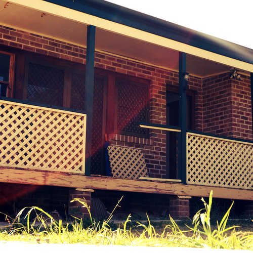 A brick house with a front porch featuring lattice fencing, steps, and a grassy area in the foreground stands as a reminder amidst the ongoing home demolitions in the neighborhood.