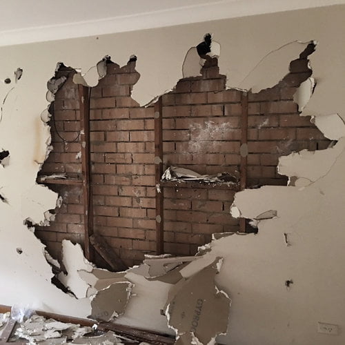 A large section of drywall is broken, revealing brickwork and wooden studs behind the damaged wall. Debris, typical of home demolitions, is scattered on the floor below.