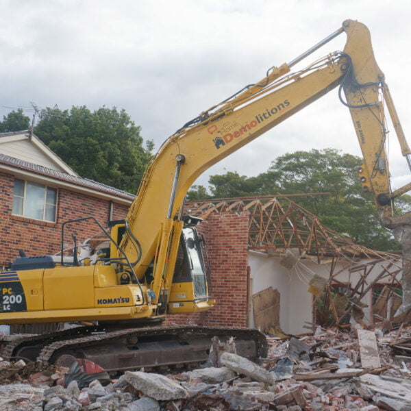West Ryde, NSW 2214 5 A yellow excavator engaged in home demolitions, tearing down a brick house surrounded by rubble and debris, with a partially collapsed roof visible in the background.