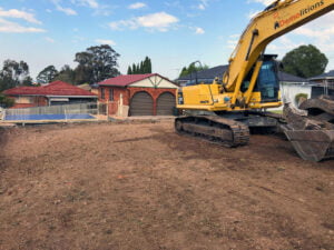 A yellow excavator is parked on a freshly cleared plot of land in front of residential houses, likely after recent home demolitions. The ground appears to be leveled and prepared for construction.