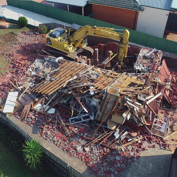 Greystanes, NSW 2145 3 A bulldozer sits in the middle of a demolished house, surrounded by piles of bricks, wood, and debris from recent home demolitions. Nearby buildings and a grassy area are visible in the background.