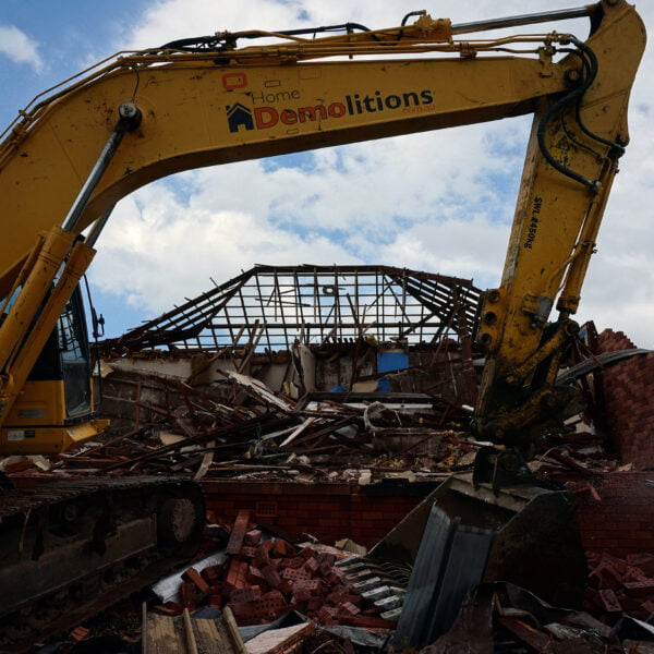 A yellow excavator from Home Demolitions demolishes a house, with rubble and debris scattered around. The roof structure is partially collapsed.