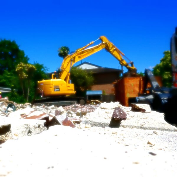 A yellow excavator is seen performing home demolitions, reducing a building to rubble under a clear blue sky.